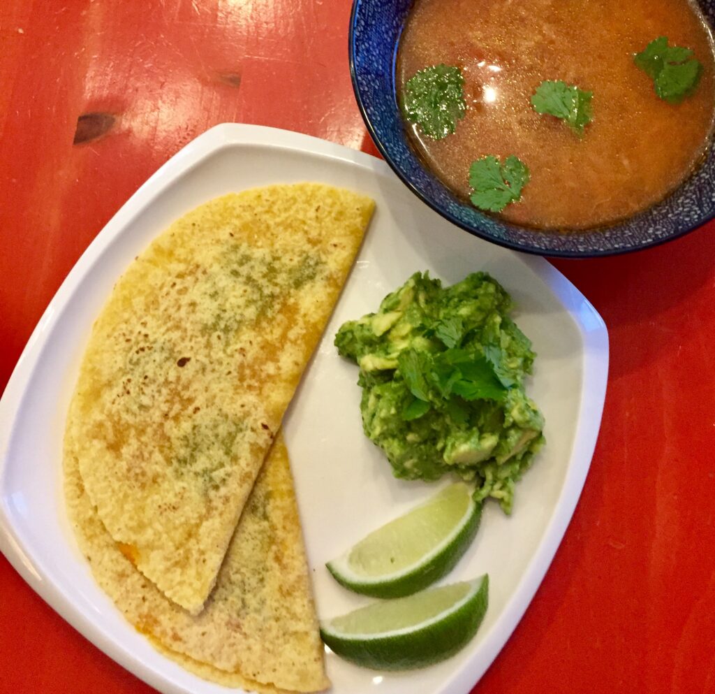 A blue bowl filled with red fideo soup and garnished with green chopped cilantro sits above and to the right of a large white plate filled with two cheese quesadillas and a serving of guacamole garnished with lime wedges. The table underneath is red.